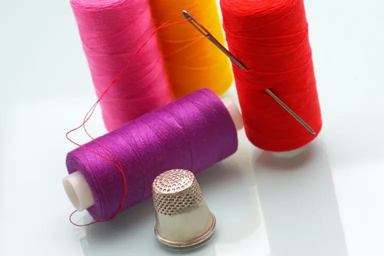 Close-up of colorful thread spools in pink, purple, orange, and red with a metal thimble and sewing needle on a white surface, illustrating sewing tools used in fabric construction.