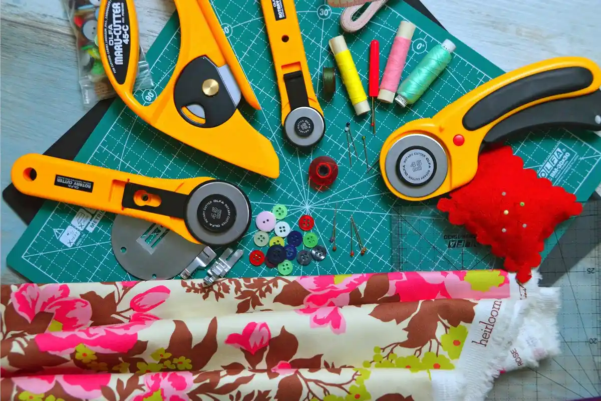 A top-down view of a sewing and fabric repair setup on a green cutting mat, featuring yellow rotary cutters, thread spools, a red pincushion, buttons, and folded floral fabric.