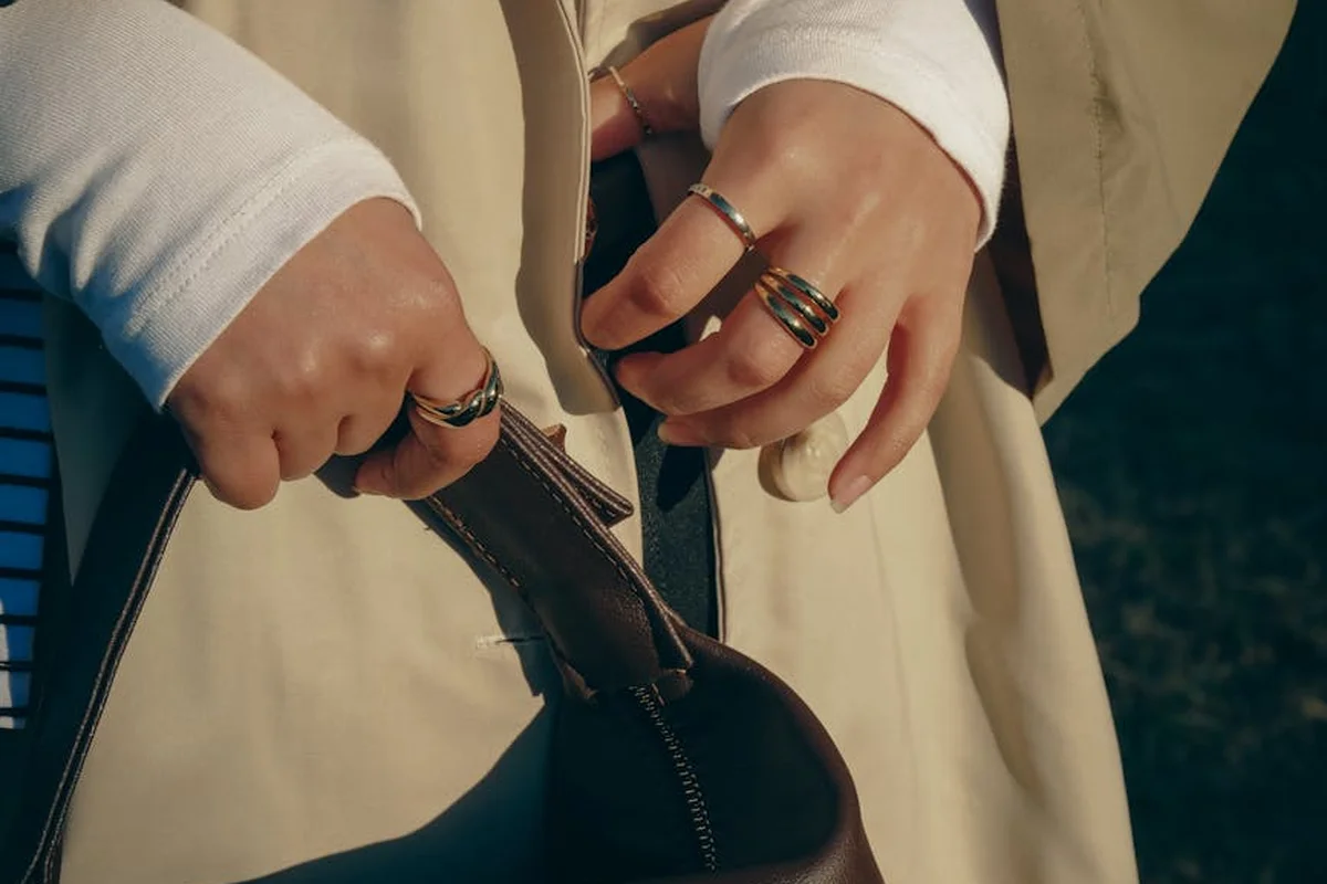 Close-up of hands with rings adjusting a brown leather handbag, outdoor background