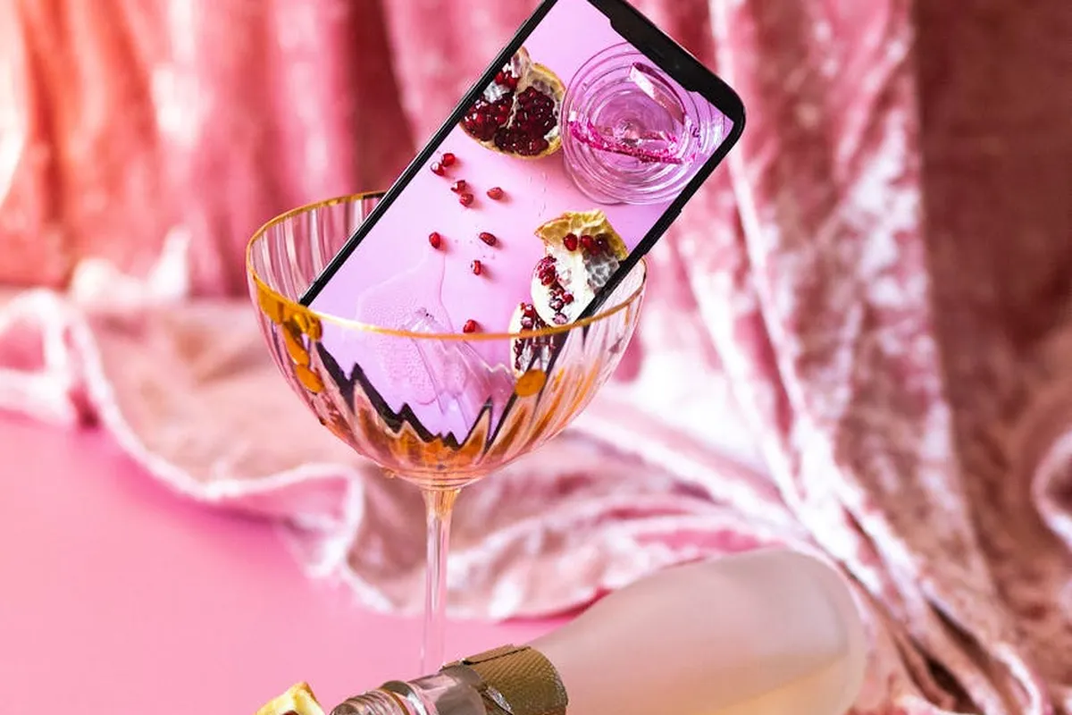 Pink velvet fabric backdrop with a tilted smartphone placed inside a champagne glass and a bottle nearby, illustrating a delicate setting related to cleaning challenges.