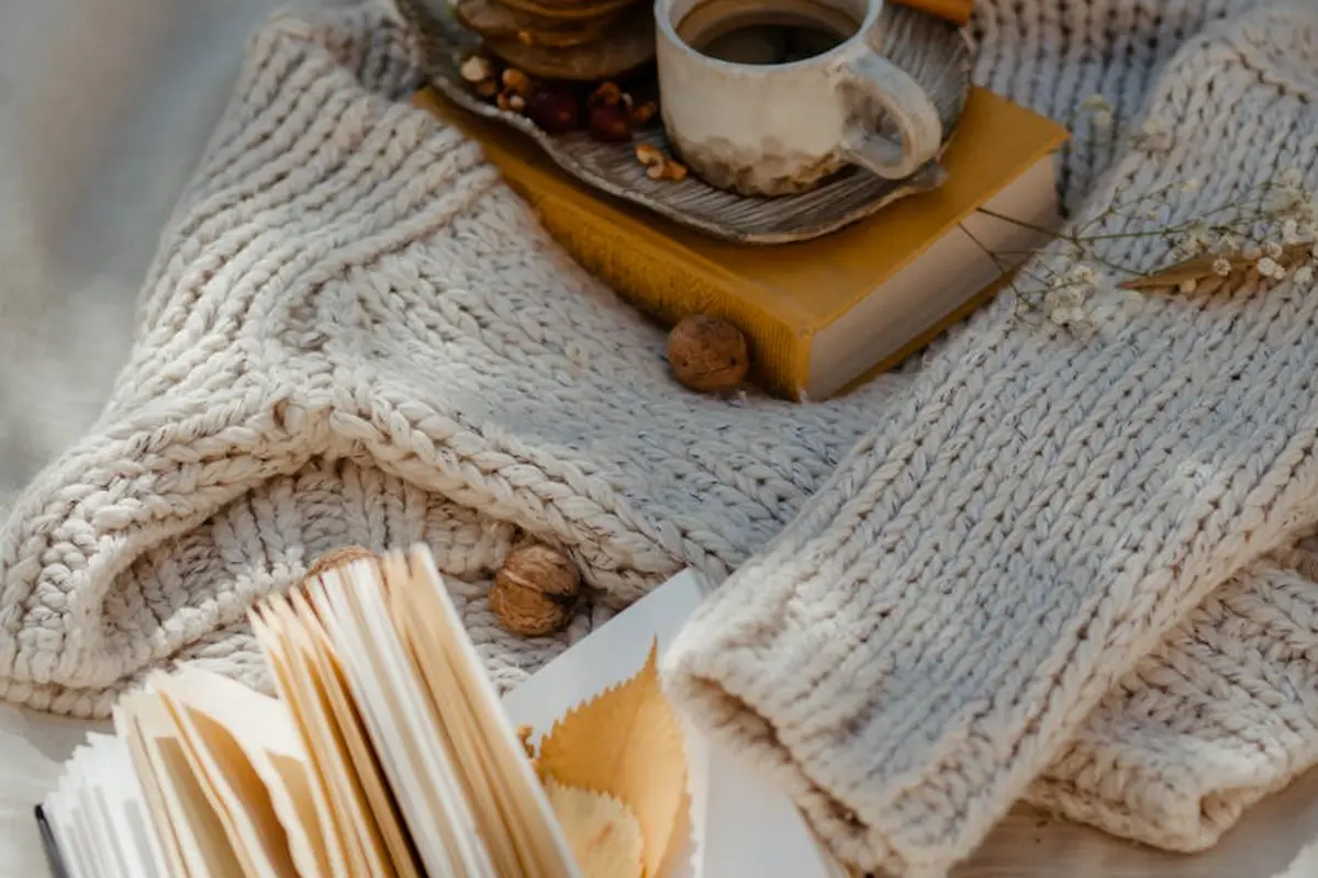 Cream chunky knit sweater with an open book, a cup on a saucer, and scattered walnuts on a wooden surface.