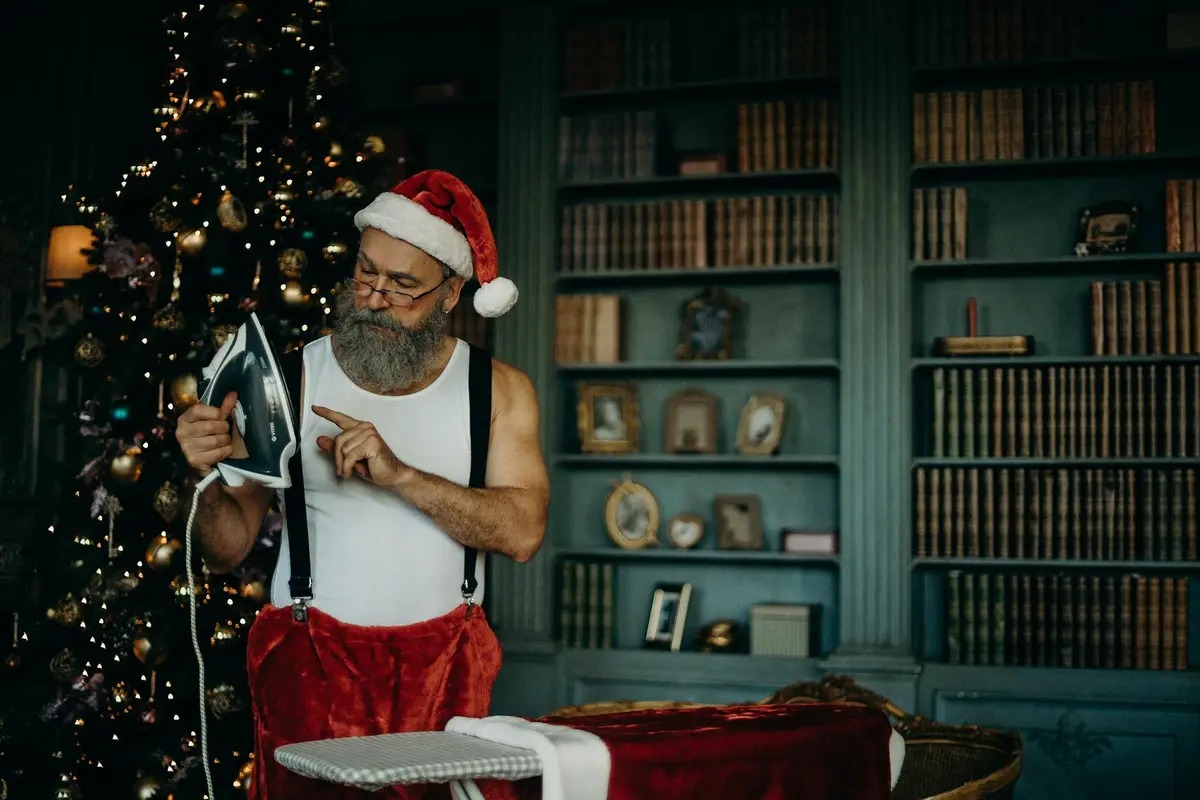 A Santa Claus figure wearing a white tank top and red suspenders irons fabric beside a decorated Christmas tree in a study lined with bookshelves.