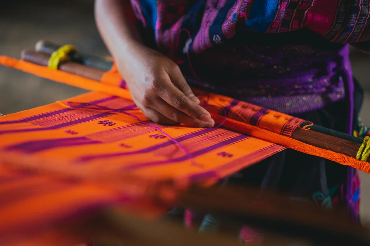 Close-up of hands weaving bright orange fabric with purple patterns on a loom