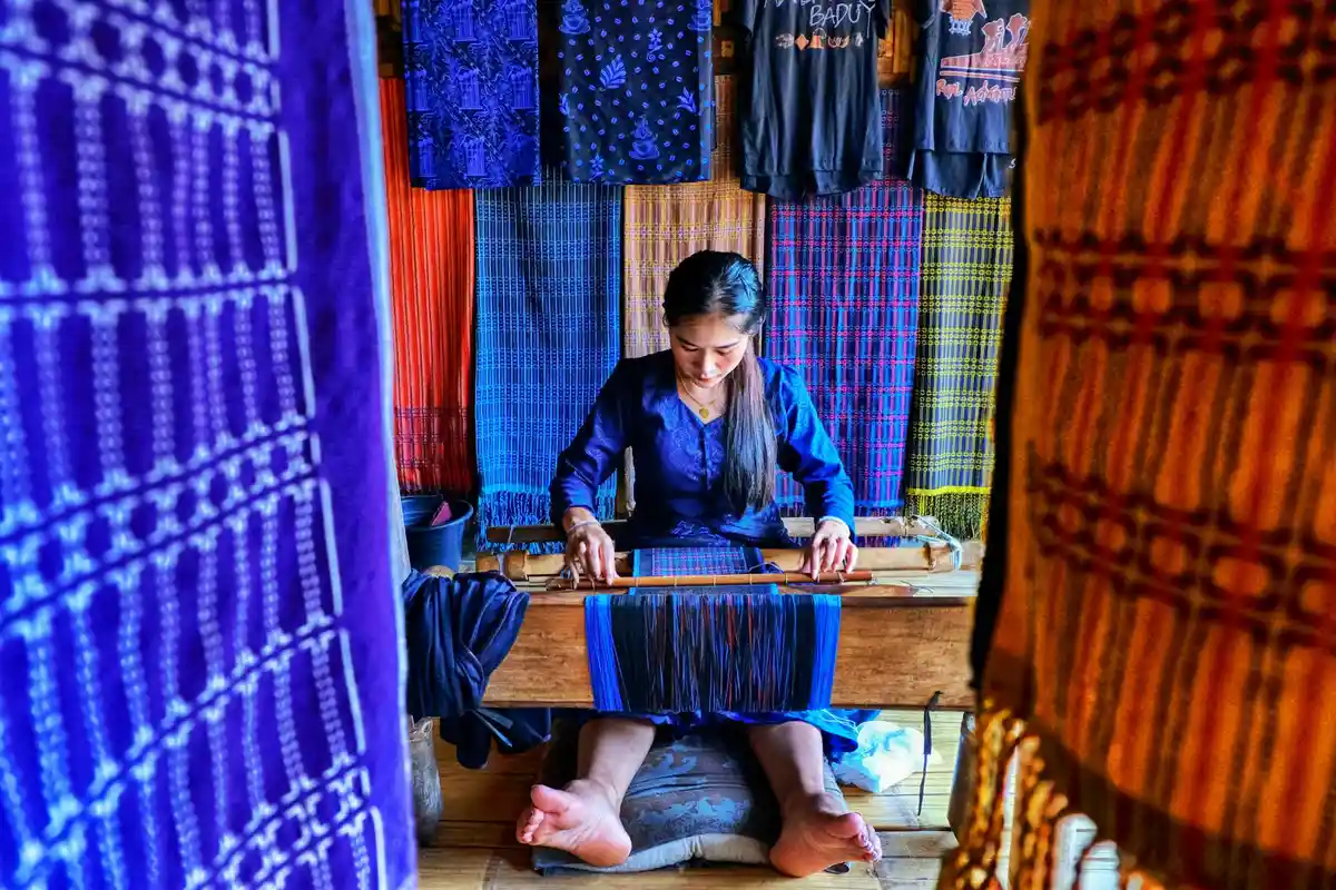 A person seated at a wooden loom weaving blue fabric, surrounded by vibrant hanging textiles in purple, blue, and orange.