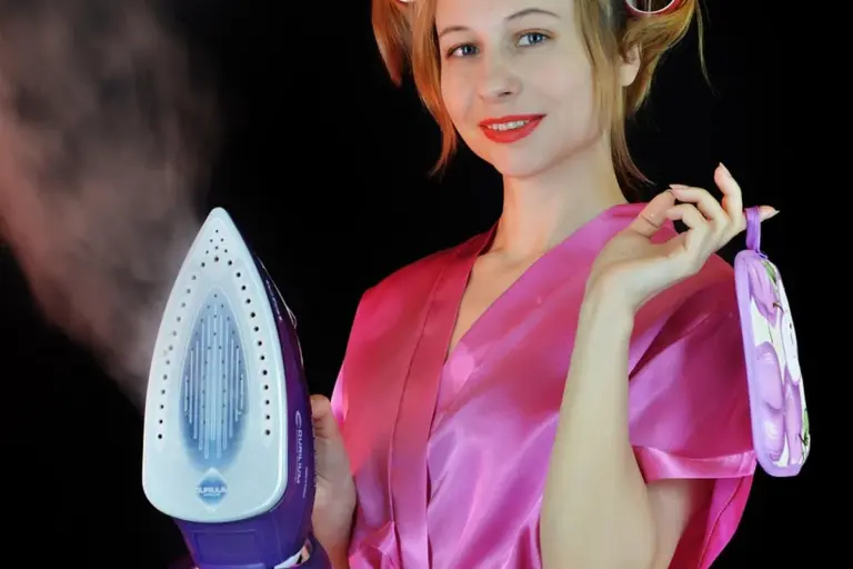 Smiling woman in a pink satin robe irons fabric with a blue and white steam iron, steam rising from the iron.