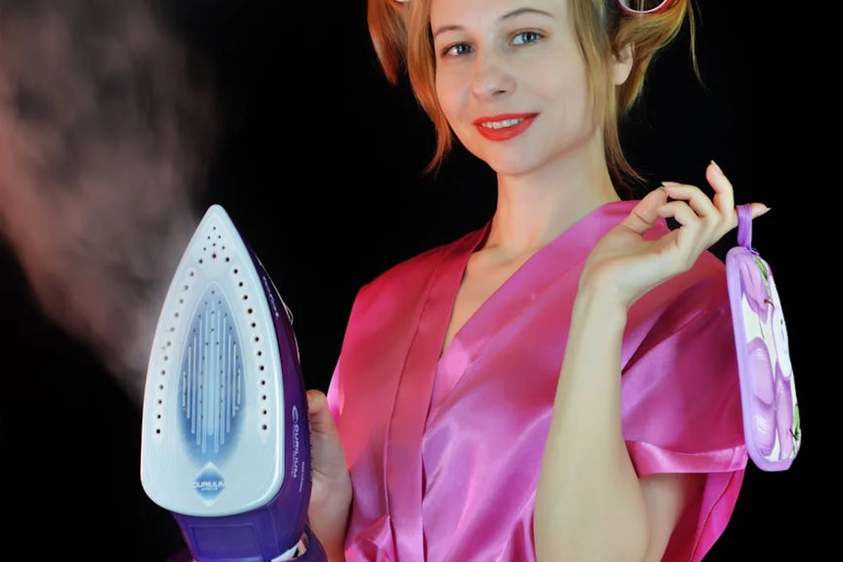 A person in a pink satin robe irons a garment with a blue and white steam iron, steam rising around the iron.